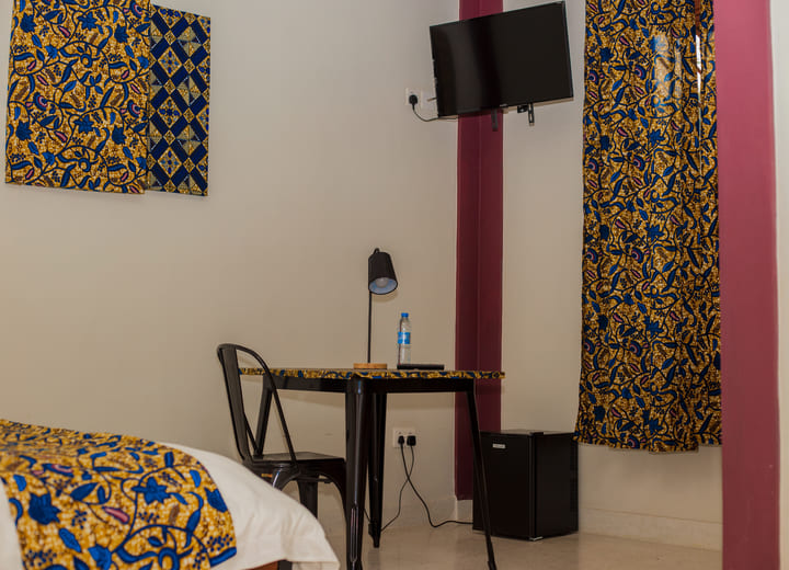 A well-furnished hotel room with traditional African-themed decor, featuring a workspace with a table, chair, and desk lamp. A wall-mounted TV is present above a mini-fridge, and the room includes vibrant patterned curtains and bedding.