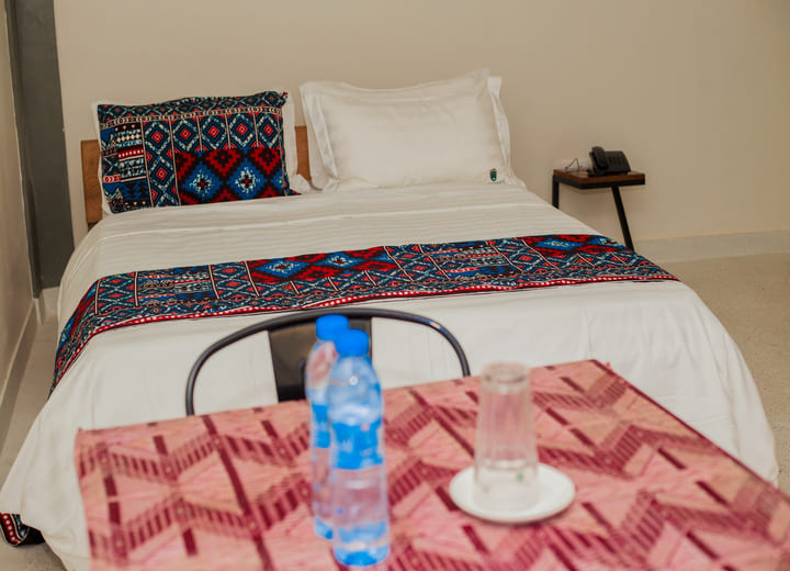 A cozy hotel room with a neatly made bed featuring vibrant African-patterned decor, a bedside table with a phone, and a dining table with bottled water and a glass.
